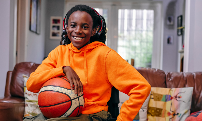 Smiling child in an orange hoodie and headphones, holding a basketball, sitting in a cozy living room.